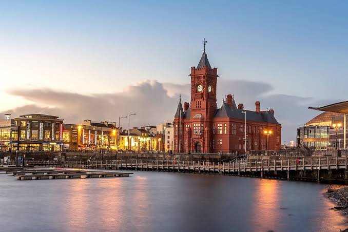 A red-brick clock tower on a British harbour at dusk, its lights reflected in still water.